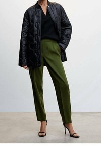 Veste noire matelassée en tissu texturé, associée à un pantalon olive ajusté. Le modèle porte des talons aiguilles noirs, créant une silhouette moderne.