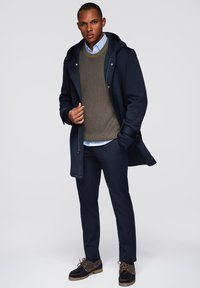 Abrigo navy con capucha sobre un suéter de punto oliva, camisa azul clara y pantalones oscuros. Zapatos marrones con cordones y suelas texturizadas.