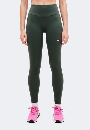 Modèle portant un legging Nike vert foncé à taille haute et des chaussures de course rose vif, sur un fond uni.