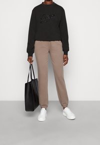 Czarna bluza z cekinowym logo "Guess", beżowe spodnie dresowe, białe sneakersy i czarna torba na ramię, stojąca na jasnoszarym tle.