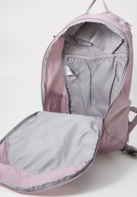 Sac à dos rose clair avec doublure grise, présentant un compartiment principal ouvert et une poche avant. Le matériau est en tissu, affichant une texture lisse.