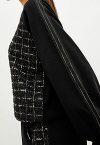 Top in bianco e nero con texture, caratterizzato da un design intrecciato a scacchi e maniche in maglia nera con un sottile dettaglio a strisce metalliche.