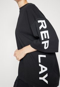 Černé oversized tričko vyrobené z bavlny, se silným bílým nápisem "REPLAY" na levém rukávu, s kulatým výstřihem a krátkými rukávy.