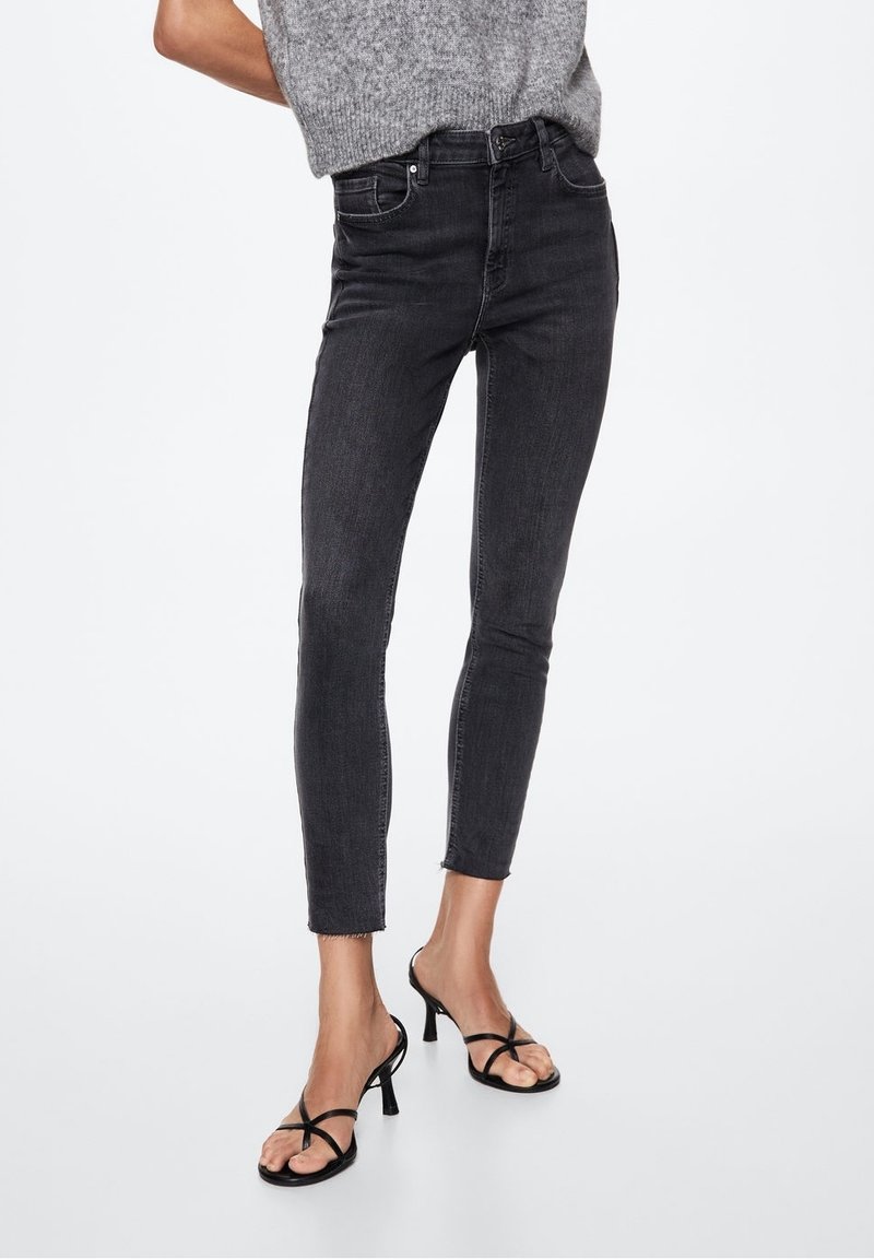Personne portant un jean skinny noir taille haute avec des ourlets effilochés et des sandales à talons noires à brides sur fond blanc.