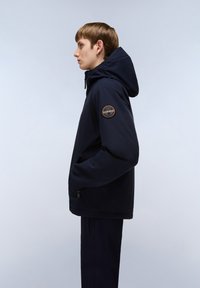 Chaqueta softshell navy con capucha, que incluye un bolsillo lateral con cremallera y un parche de logo circular en la manga izquierda. Textura suave, diseño moderno.
