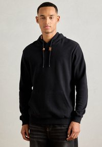 Junger Mann steht vor einem neutralen Hintergrund und trägt einen schwarzen Hoodie mit einer Fronttasche und dunklen Jeans.
