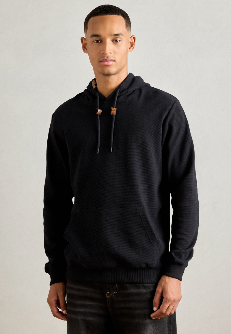 Junger Mann steht vor einem neutralen Hintergrund und trägt einen schwarzen Hoodie mit einer Fronttasche und dunklen Jeans.