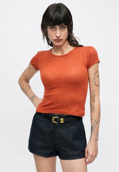 Τοπ με κοντά μανίκια σε σκουριά πορτοκαλί απόχρωση με ραβδωτή υφή, στρογγυλή λαιμόκοψη, φορεμένο με σκούρα τζιν σορτς και μαύρη ζώνη με χρυσή αγκράφα.