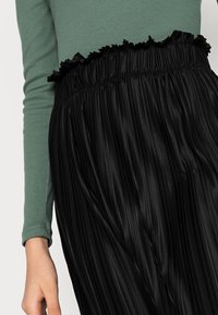 Jupe plissée noire avec une ceinture froncée, fabriquée en tissu brillant. Un bras portant un haut ajusté vert est partiellement visible dans le cadre.