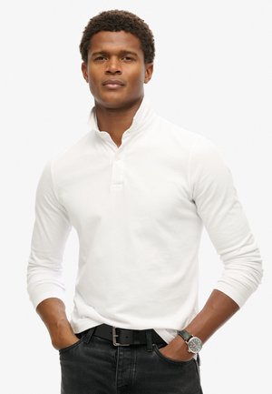 Weißes langärmliges Polohemd mit Knopfleiste und erhöhtem Kragen. Das Modell trägt einen schwarzen Gürtel und eine runde Uhr mit einem dunklen Armband.