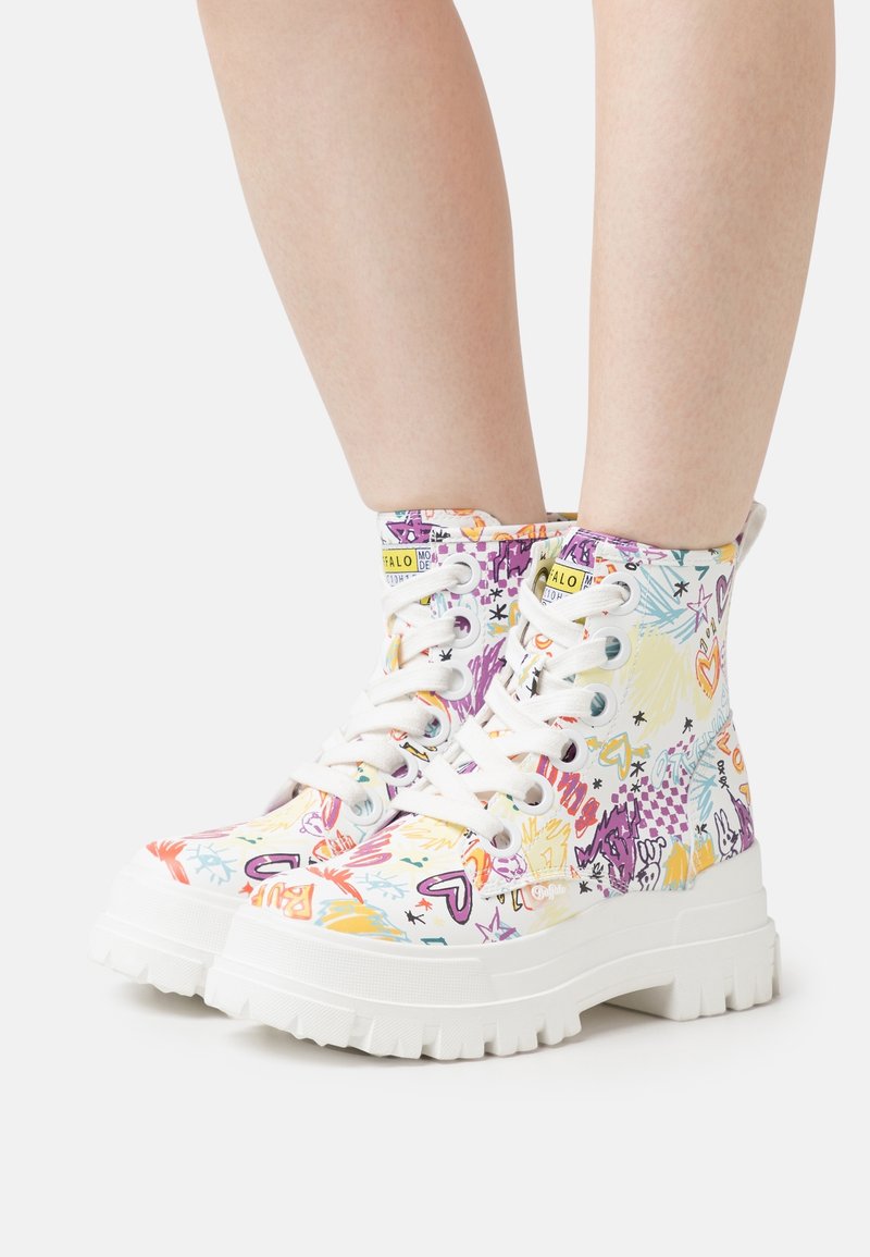 Personne portant des baskets montantes à plateforme blanches avec des motifs colorés de style graffiti, lacets attachés, debout sur un fond blanc.