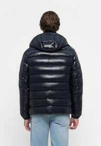Moški nosi sijoč temno moder puffer jopič s kapuco in svetlo modre kavbojke, prikazan od zadaj na enobarvni sivi podlagi.