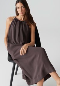 Langes, ärmelloses Kleid aus dunkelbraunem Stoff, mit einem gerafften Ausschnitt und lockerer Passform, das auf einem schwarzen Stuhl ruht. Glatte Textur.