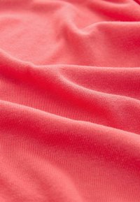 Korallfarbener Stoff mit glatter Textur, der feine Rippungen und sanfte Falten zeigt. Das Material wirkt weich und leicht, ideal für lässige Kleidung.
