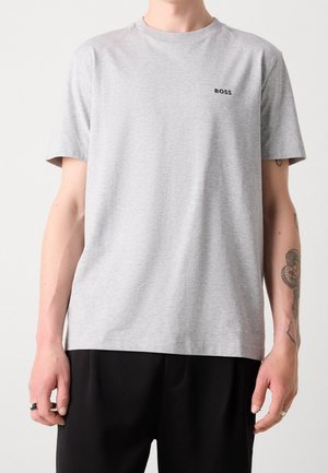 Man draagt een lichtgrijs T-shirt met korte mouwen met het logo "BOSS" op de borst en een zwarte broek, toont een getatoeëerde rechter onderarm en ringen aan de vingers.