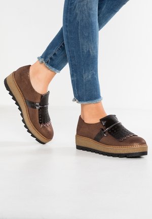 Brune ruskindssko med sorte lakerede detaljer og dekorative perforeringer. Har en tekstureret sål med en let platformshøjde.