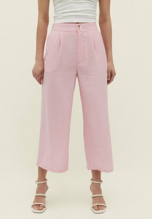 Kobieta nosząca jasnoróżowe, wysokowalne, szerokie spodnie 7/8 z zapięciem na guziki, w połączeniu z białymi sandałami na obcasie i białym topem bez rękawów.