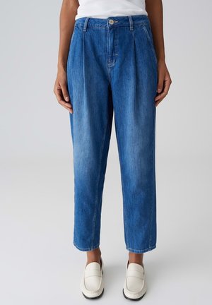 Femme portant un jean en denim bleu taille haute et coupe décontractée, associé à des mocassins blancs sans lacets, debout devant un fond uni.