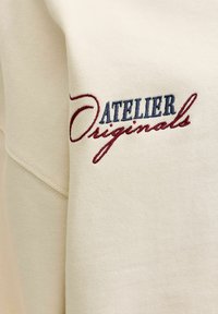 Υφασμάτινο κρεμ με κεντημένο κείμενο "ATELIER Originals" σε μπλε και μαρόν νήμα σε ρούχο.