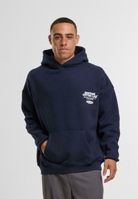 Tamsiai mėlynas medvilninis džemperis su kengūros kišene, dideliu gobtuvu, baltu tekstu ant krūtinės ir renkamu rankovėse bei apačioje.