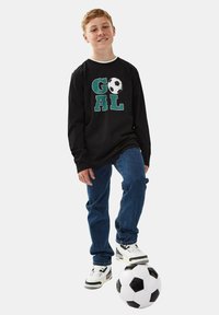Schwarzes Langarmshirt mit grünem und weißem "GOAL"-Text und Fußballgrafik, kombiniert mit blauen Jeans und schwarz-weißen Sneakern.