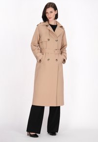 Trench coat bej cu talie centurată, cu un front cu două rânduri de butoane și butoane mari. Are un guler clasic și mâneci lungi.