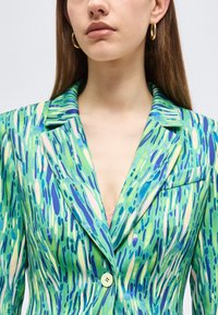 Jeune femme portant un blazer à motifs verts, bleus et crème avec des boucles d'oreilles en forme d'anneaux dorés, montrant le décolleté et une partie d'un haut rose en-dessous.