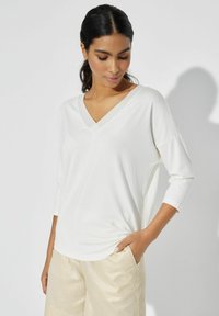 Biała, lekka bluzka z dekoltem w serek i rękawami 3/4. Miękka faktura, luźny krój. Noszona z szortami z jasnego, błyszczącego materiału.