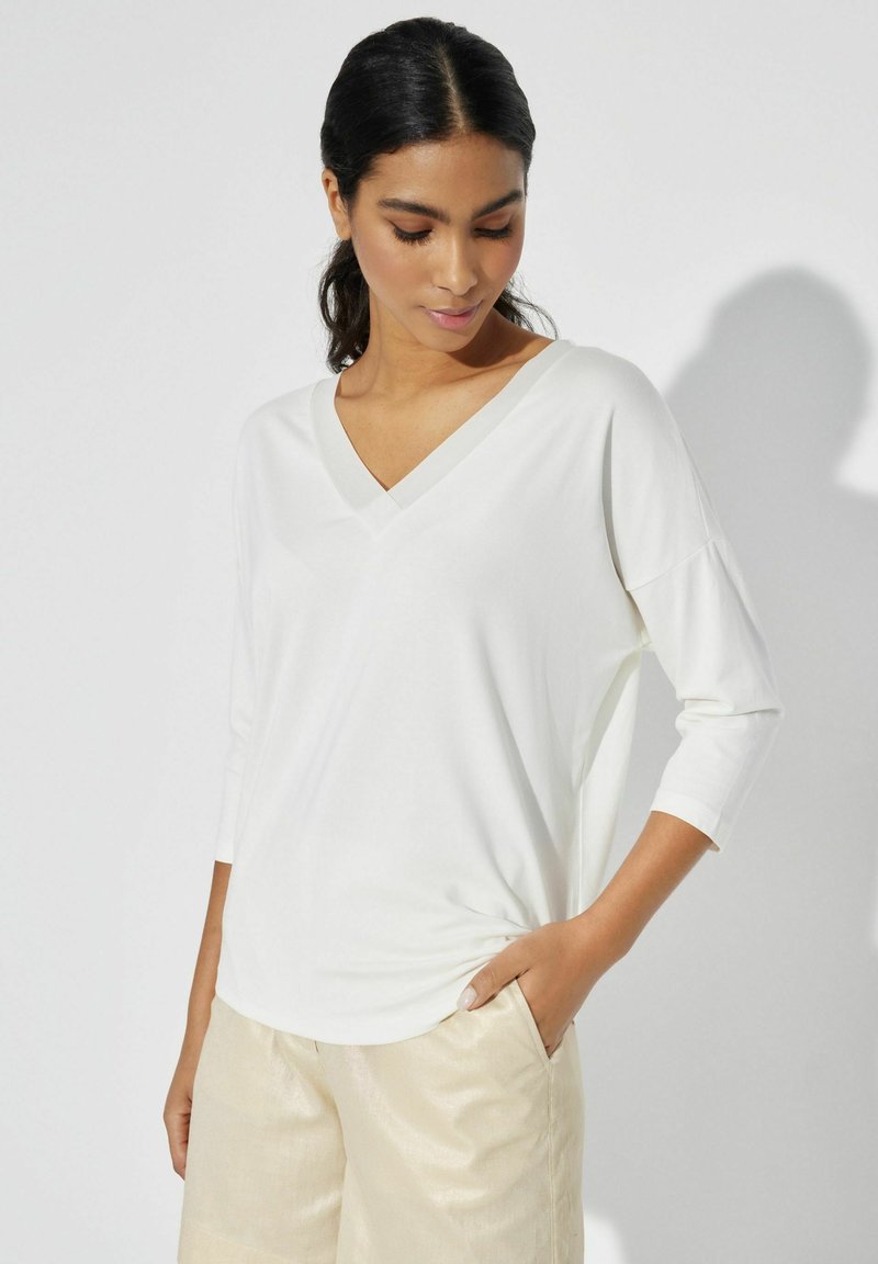 Biała, lekka bluzka z dekoltem w serek i rękawami 3/4. Miękka faktura, luźny krój. Noszona z szortami z jasnego, błyszczącego materiału.