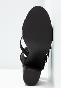 Semelle de sandale noire à talon, vue de face, montrant le dessous texturé et le talon bloc avec des brides à la cheville et au pied.