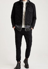 Zwarte denimjack over een witte ribbeltrui, gecombineerd met zwarte cargobroek. De outfit bevat enkellaarsjes en een geruit flanellen shirt.
