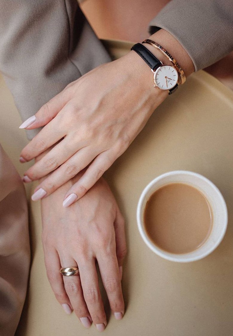 Zwei Hände mit hellrosa Nägeln, eine trägt einen goldenen Ring und die andere eine Uhr mit schwarzem Armband, die neben einer Tasse Kaffee ruhen.