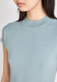 Šviesiai mėlyna trumpomis rankovėmis viršutinė dalis su aukštu kaklu ir vagoti tekstūra, su mažuoju baltu "ck" logotipu kairėje pusėje.