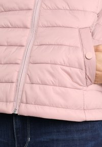 Chaqueta rosa acolchada con cierre de cremallera y un bolsillo con botón a presión. Presenta líneas de acolchado horizontal y una textura de tela suave y brillante.