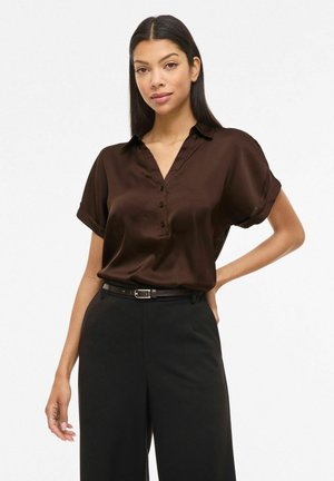 Femme aux longs cheveux foncés portant un chemisier marron foncé à manches courtes boutonné, rentré dans un pantalon noir taille haute avec ceinture.