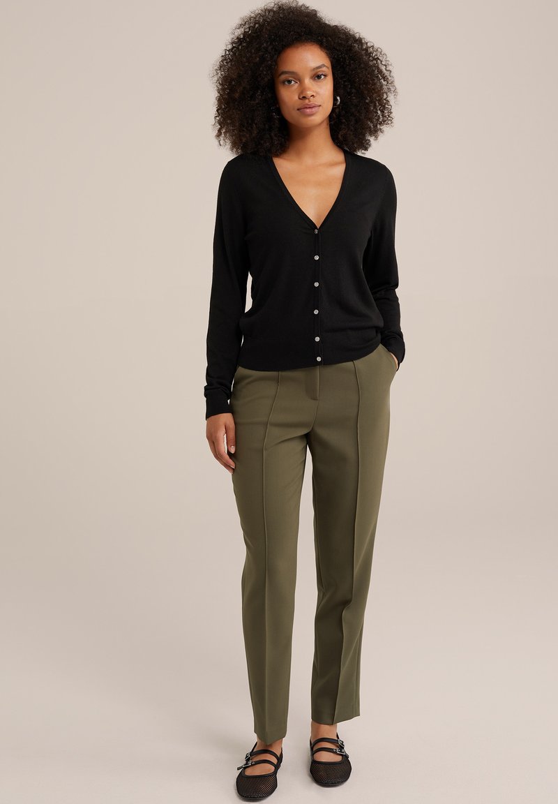 Zwarte button-up cardigan met een V-hals, gecombineerd met olijfgroene tailored broek met plooien. Zwarte mesh ballerina's maken de outfit af.