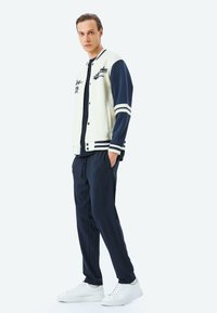 Chaqueta de varsity para hombres en crema con mangas azul marino, con acentos a rayas y detalles bordados. Combinada con pantalones jogger azul marino y zapatillas blancas.