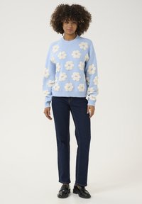 Gai zils džemperis ar krēmveida ziedu applikācijām, apaļš kakls, ribota piedurknes; kombinēts ar tumši ziliem džinsiem un melniem mokasīniem.