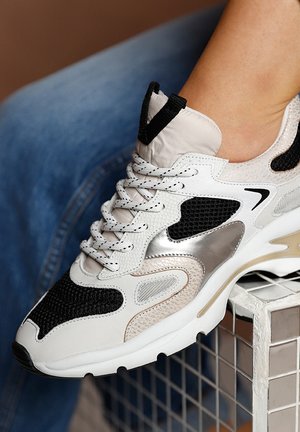 De sneaker heeft een mix van witte, zwarte en beige materialen, met gaas- en glanzende accenten, een textiel bovenwerk en een stevige zool.