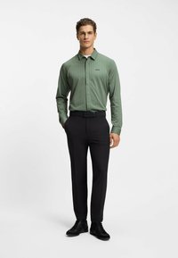 Camicia a maniche lunghe verde a fantasia con colletto a punta, abbinata a pantaloni neri e sneakers nere. La camicia presenta un logo sul petto.