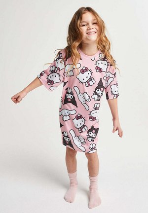 Junges Mädchen lächelt, trägt ein pinkfarbenes, übergroßes Kleid, das mit Hello Kitty und Cartoon-Charakteren bedruckt ist, steht barfuß vor weißem Hintergrund.