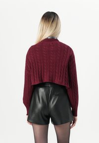Frau mit langen Ombre-Haaren, die einen burgunderfarbenen, kurz geschnittenen Pullover aus Kabelstrick, schwarze Lederhosen und transparente schwarze Strumpfhosen trägt, mit dem Rücken zugewandt.