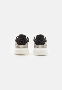 Baskets blanches avec un talon en tissu noir texturé, ornées de petites perles. Semelle blanche lisse, couture renforcée à l'arrière.