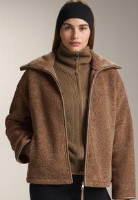 Hnědá umělá kožešinová bunda se zipem vpředu, oversized límcem a hnědým žebrovaným rolákem. Textura je měkká, s kontrastními hnědými akcenty.