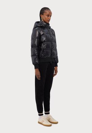 Čierna, lesklá prešívaná bunda s kapucňou, so zipsom vpredu a elastickými manžetami, skombinovaná s čiernymi nohavicami a krémovými teniskami s červenými pruhmi.