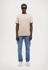 T-shirt beige in cotone a maniche corte, jeans di denim blu chiaro e sneakers bianche. Accessoriato con occhiali da sole neri.