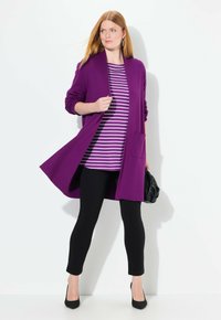 Lila offene Strickjacke mit langen Ärmeln, getragen über einem gestreiften lila-weißen Hemd, schwarzen Leggings und schwarzen High Heels.