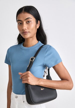 Mujer con pelo largo y negro lleva una camiseta azul de manga corta y pantalones blancos, sosteniendo un bolso de cuero negro con hebilla dorada.