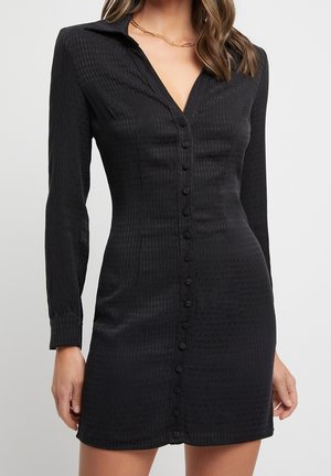 Femme portant une robe noire ajustée à manches longues, avec des boutons recouverts de tissu à l'avant et un col, mettant en valeur la taille et les hanches.
