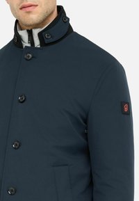 Cappotto isolato blu navy con colletto alto, rifiniture nere e numerosi bottoni. Presenta una tasca laterale e una toppa con il marchio sulla manica.